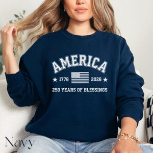 Pu&ograve; includere: Felpa blu navy con la scritta "AMERICA" in lettere bianche. Sotto, una bandiera americana, gli anni 1776 e 2026 e la scritta "250 YEARS OF BLESSINGS".