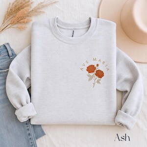 Peut inclure: Un sweat-shirt gris clair avec les mots "AVE MARIA" dans un design circulaire, accompagn&eacute; d'une illustration florale de deux roses orange. Le mot "Ash" est imprim&eacute; en bas de l'image.