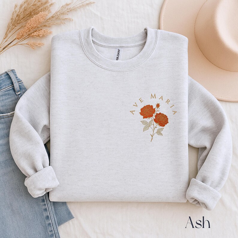 Peut inclure: Un sweat-shirt gris clair avec les mots "AVE MARIA" dans un design circulaire, accompagn&eacute; d'une illustration florale de deux roses orange. Le mot "Ash" est imprim&eacute; en bas de l'image.