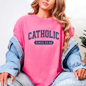 May include: A bright pink sweatshirt with the word "CATHOLIC" in navy blue arched lettering. Below the word is a navy blue rectangle with the text "SINCE 33 AD" in white. The sweatshirt is part of a casual clothing collection.