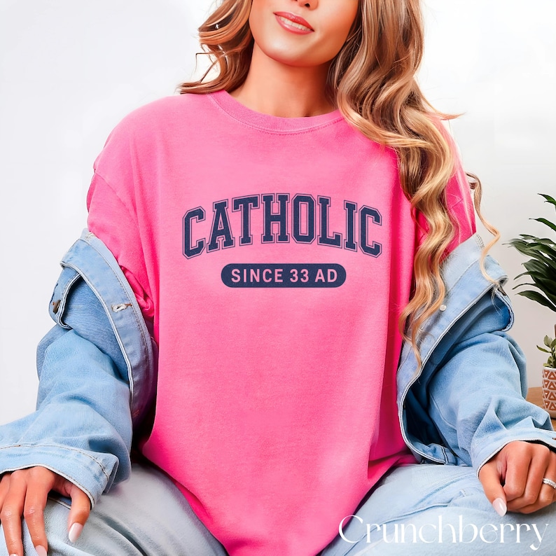 May include: A bright pink sweatshirt with the word "CATHOLIC" in navy blue arched lettering. Below the word is a navy blue rectangle with the text "SINCE 33 AD" in white. The sweatshirt is part of a casual clothing collection.