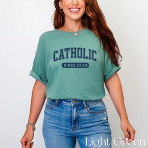 May include: A light green t-shirt with the word "CATHOLIC" arched across the chest in navy blue block letters, with "SINCE 33 AD" below. The shirt is paired with blue jeans.