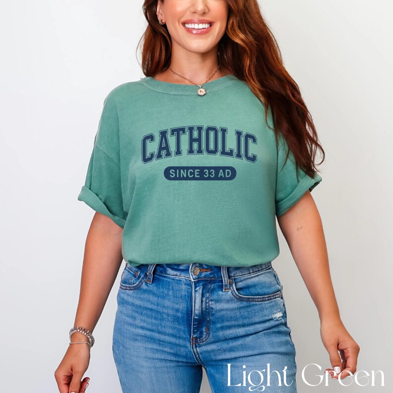 May include: A light green t-shirt with the word "CATHOLIC" arched across the chest in navy blue block letters, with "SINCE 33 AD" below. The shirt is paired with blue jeans.