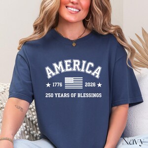 Pu&ograve; includere: T-shirt blu navy con la scritta "AMERICA" in lettere bianche sopra una grafica della bandiera americana. Sotto la bandiera ci sono i numeri "1776" e "2026" con stelle su entrambi i lati e il testo "250 YEARS OF BLESSINGS".