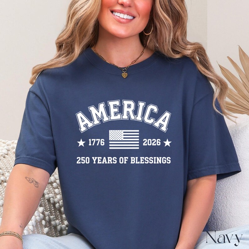 Pu&ograve; includere: T-shirt blu navy con la scritta "AMERICA" in lettere bianche sopra una grafica della bandiera americana. Sotto la bandiera ci sono i numeri "1776" e "2026" con stelle su entrambi i lati e il testo "250 YEARS OF BLESSINGS".