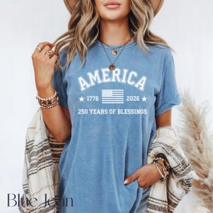 Pu&ograve; includere: Una maglietta blu con la scritta "AMERICA" in lettere bianche arcuate. Sotto la parola "AMERICA" c'&egrave; una grafica bianca della bandiera americana, con gli anni "1776" e "2026" su entrambi i lati. Il testo "250 YEARS OF BLESSINGS" &egrave; sotto la bandiera.