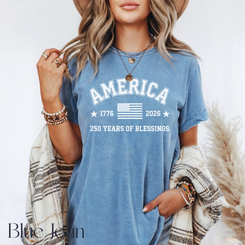 Pu&ograve; includere: Una maglietta blu con la scritta "AMERICA" in lettere bianche arcuate. Sotto la parola "AMERICA" c'&egrave; una grafica bianca della bandiera americana, con gli anni "1776" e "2026" su entrambi i lati. Il testo "250 YEARS OF BLESSINGS" &egrave; sotto la bandiera.