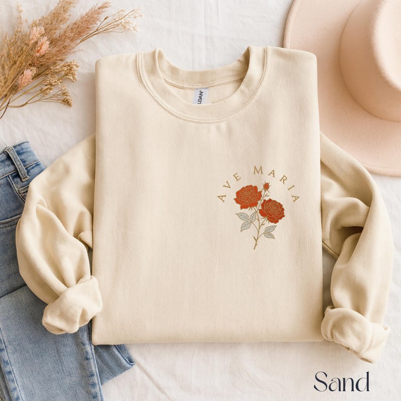 Peut inclure: Sweat-shirt couleur sable avec l'inscription "AVE MARIA" et un motif floral de deux roses rouges. Le sweat-shirt &agrave; col rond et &agrave; manches longues est pli&eacute; proprement. Le mot "Sand" est imprim&eacute; en bas de l'image.