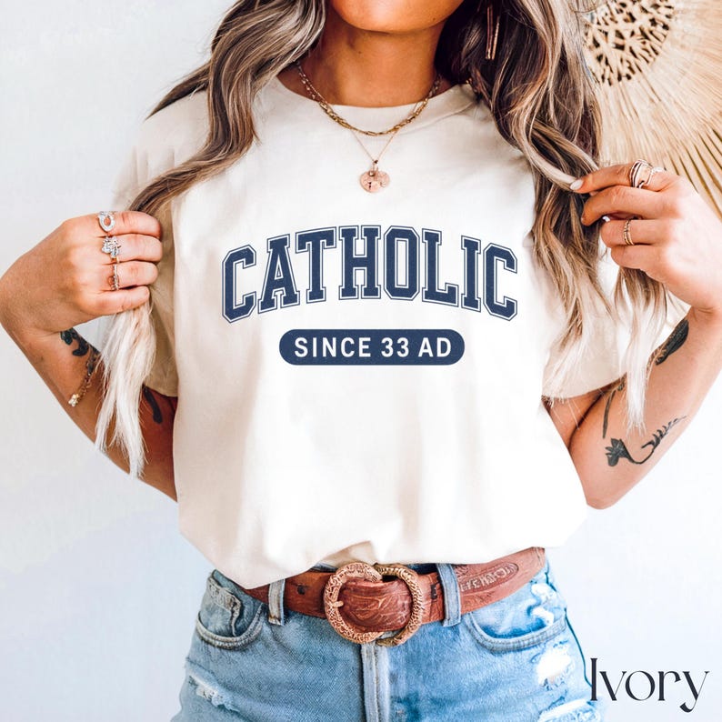 May include: Ivory-colored t-shirt with the word "CATHOLIC" in navy blue block letters, arched across the chest. Below, a navy blue rectangular banner reads "SINCE 33 AD". The shirt is paired with blue jeans and a brown belt.