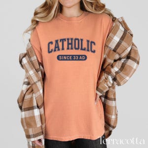 May include: Terracotta-colored t-shirt with the word "CATHOLIC" in navy blue arched lettering. Below the word is the text "SINCE 33 AD" in a navy blue oval. The shirt is worn with a brown and tan plaid flannel.