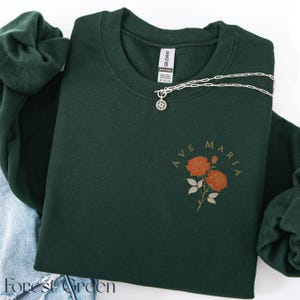 Peut inclure: Sweat-shirt vert for&ecirc;t avec l'inscription "AVE MARIA" et un motif floral de roses orange. Un collier en argent avec un pendentif en forme de soleil est drap&eacute; sur le sweat-shirt. Le sweat-shirt est pli&eacute; avec un v&ecirc;tement en jean et un autre v&ecirc;tement vert.