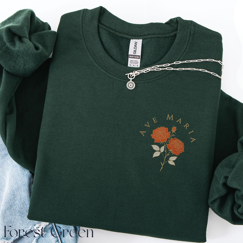 Peut inclure: Sweat-shirt vert for&ecirc;t avec l'inscription "AVE MARIA" et un motif floral de roses orange. Un collier en argent avec un pendentif en forme de soleil est drap&eacute; sur le sweat-shirt. Le sweat-shirt est pli&eacute; avec un v&ecirc;tement en jean et un autre v&ecirc;tement vert.