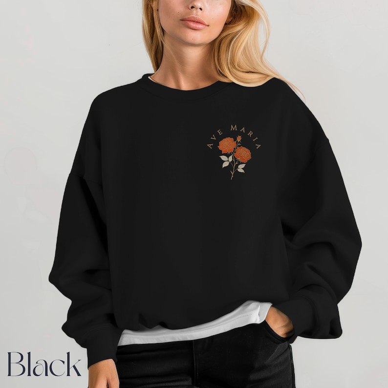 Peut inclure: Sweat-shirt noir avec le texte "AVE MARIA" et un motif floral orange. Le sweat-shirt &agrave; col rond a des manches longues. Le mot "Black" est imprim&eacute; en bas &agrave; gauche de l'image.