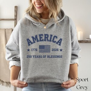 Pu&ograve; includere: Felpa con cappuccio grigio sportivo con la scritta "AMERICA" in blu navy ad arco sul petto. Sotto, una bandiera americana blu navy, gli anni "1776" e "2026" e la frase "250 YEARS OF BLESSINGS".