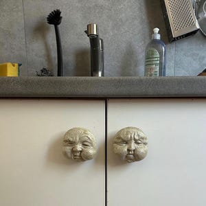 K&ouml;nnte beinhalten: Zwei Schrankkn&ouml;pfe mit einem geformten Gesichtsdesign. Die Kn&ouml;pfe sind hellbeige und haben eine runde Form. Die Kn&ouml;pfe sind an einer wei&szlig;en Schrankt&uuml;r befestigt.