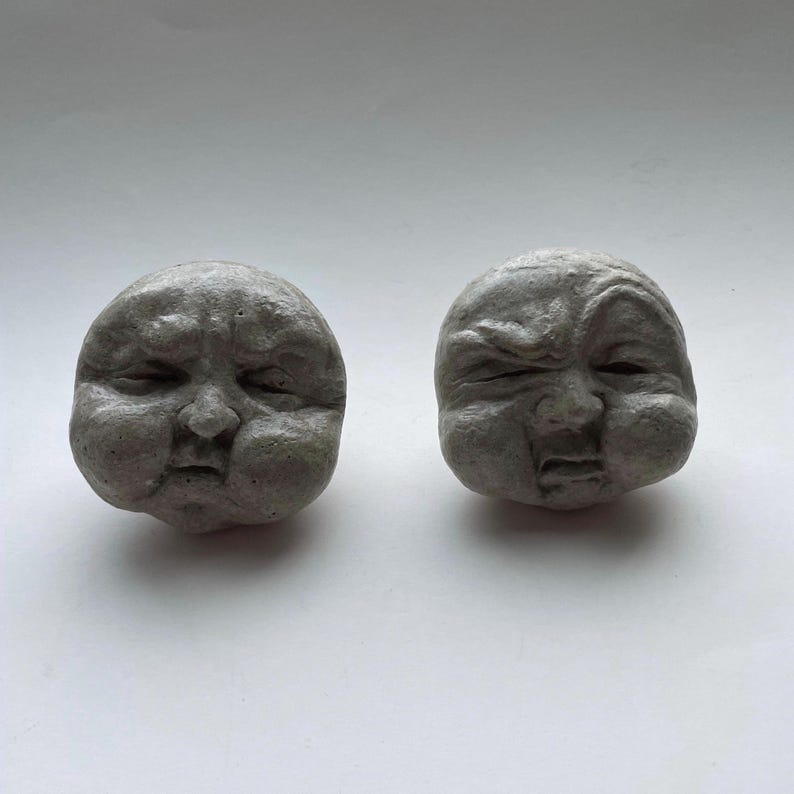 K&ouml;nnte beinhalten: Zwei graue, steinartige Skulpturen von Babygesichtern. Ein Gesicht hat geschlossene Augen, das andere ein zusammengekniffenes Auge und einen leicht ge&ouml;ffneten Mund. Die Skulpturen haben eine runde Form und eine strukturierte Oberfl&auml;che mit sichtbaren Gesichtsz&uuml;gen.