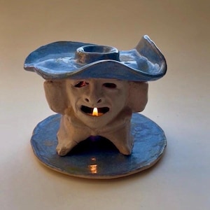 Pode incluir: Um castiçal de cerâmica em forma de rosto usando um chapéu azul. O rosto é esbranquiçado com a boca bem aberta e uma vela acesa dentro. O castiçal assenta sobre uma base circular azul.