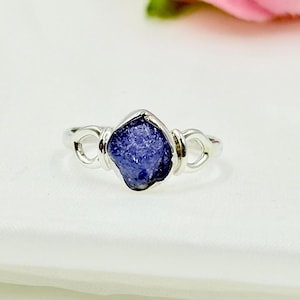 Könnte beinhalten: Ein Silberring mit einem rohen, tiefblauen Edelstein. Der Stein ist in einer Lünettenfassung gefasst und hat auf beiden Seiten dekorative Schlaufen. Der Ring wird auf einer weißen Oberfläche präsentiert.