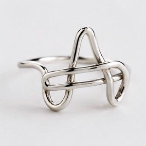 Può includere: Un anello color argento caratterizzato da un design a filo continuo che forma la lettera A. Il metallo presenta una finitura liscia e lucida con una superficie riflettente, e la struttura è composta da anelli e linee interconnesse.