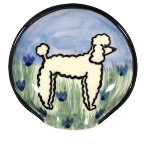 May include: A circular ceramic dish featuring a hand-painted poodle. The poodle is white with black detailing, set against a blue sky and green grass with blue flower accents. The dish has a black rim.