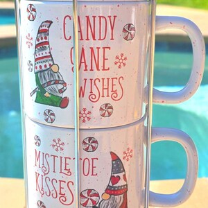 Pode incluir: Um conjunto de quatro canecas de cerâmica branca empilhadas em um suporte de metal. Cada caneca apresenta um design festivo de gnomo com bengalas doces e flocos de neve, juntamente com as frases "Mistletoe Kisses" e "Candy Cane Wishes" em vermelho.