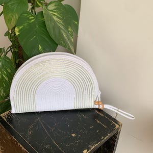 May include: White and light green woven clutch with a semi-circular design. The clutch has a zipper closure and a wrist strap with wooden beads. The bag is placed on a black wooden surface, with green leafy plants in the background.