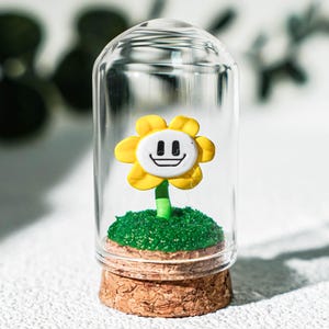 May include: A yellow flower figurine with a white face and a black smiling expression, mounted on a green textured base inside a clear glass cloche with a cork stopper base. The flower has six petals and a green stem, displayed upright.