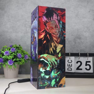 Puede incluir: Lámpara rectangular con temática de anime, con un personaje de pelo negro y puntos de sutura en la cara. La lámpara presenta una calavera y otros gráficos de estilo anime. La lámpara tiene un cable de alimentación negro y un interruptor. La lámpara está sobre una superficie blanca.