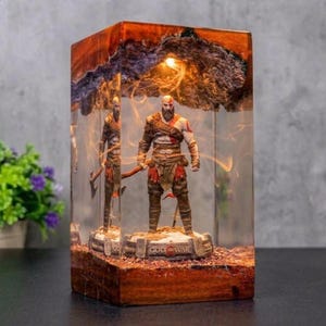 Può includere: Una lampada rettangolare in resina con una statuetta di Kratos di God of War su una base con la scritta God of War. La lampada incorpora un pezzo di legno naturale con corteccia sulla parte superiore, illuminato da una luce interna.