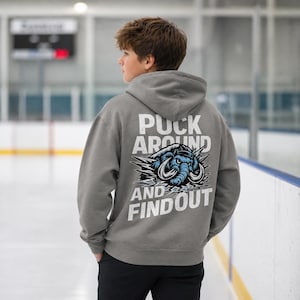 Puede incluir: Una sudadera con capucha gris con el texto "Puck Around and Find Out" y un gr&aacute;fico de elefante azul. La sudadera con capucha la lleva una persona en una pista de hielo.