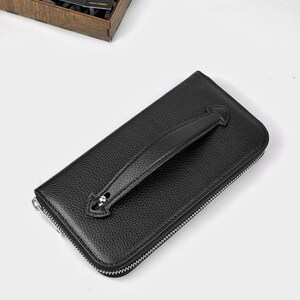 Könnte beinhalten: Eine schwarze Ledergeldbörse mit strukturierter Oberfläche und silbernem Reißverschluss. Die Geldbörse hat einen Griff und ein kleines silbernes Detail. Eine Geschenkbox mit einer "DANKE"-Karte befindet sich im Hintergrund.