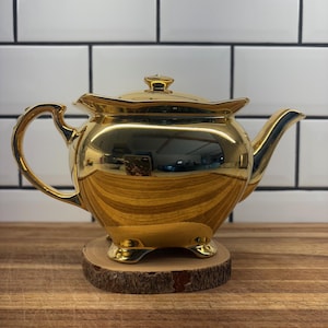 May include: A gold-coloured teapot with a curved handle and spout, placed on a small wooden slice. The teapot has a lid and rests on three small feet. The background shows a white tiled wall.