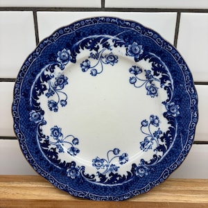 May include: A white plate with a blue floral pattern and a scalloped edge. The plate has a detailed blue border with a repeating design. The centre of the plate is white, with blue floral details.