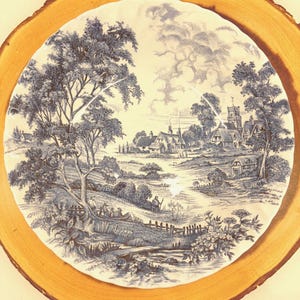 May include: A blue and white plate with a rural landscape design, presented on a circular wooden board. The plate depicts trees, buildings, and people, offering a traditional, vintage appearance. The wood slice has a diameter of approximately 30 cm.