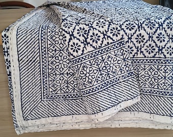 May include: A folded white and navy blue patterned quilt. The quilt features a repeating floral and geometric design. The edges are finished with a white border and stitched detailing. The quilt is folded on a wooden surface.
