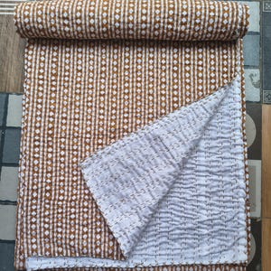 May include: A rolled-up, brown and white patterned quilt with a geometric design. The quilt is partially unfolded, revealing a white interior with stitched detailing. The quilt is made of soft material and is suitable for a bed or sofa.