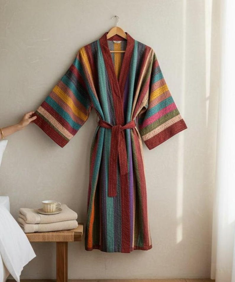 May include: A colorful, striped robe with a matching belt hangs on a wooden hanger. The robe features vertical stripes in shades of red, teal, yellow, and purple. The robe is displayed against a neutral wall, suggesting a cozy, home decor theme.