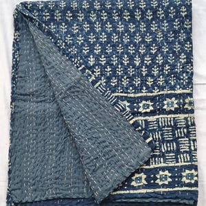 May include: An indigo blue quilt with a white floral pattern and white stitching, folded to reveal the solid blue reverse side. The quilt is a decorative home textile, made of soft fabric.