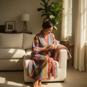 May include: A colorful, striped robe with a matching blanket draped over a white sofa. The robe features vertical stripes in shades of red, orange, purple, and teal. A woman is seated on the sofa, holding a mug. The room is filled with natural light.
