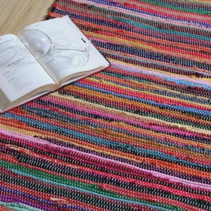 Könnte beinhalten: Ein farbenfroher, gewebter Teppich mit Streifenmuster in Rot-, Rosa-, Blau-, Grün- und Gelbtönen. Ein offenes Buch und Weingläser liegen auf dem Teppich. Der Teppich liegt auf einem hellen Holzboden.