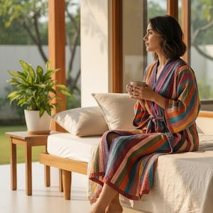 May include: A colorful striped robe with a matching belt. The robe features a V-neck and wide sleeves. The robe is made of a soft, textured fabric. The person is sitting on a bed with a cup in their hands.
