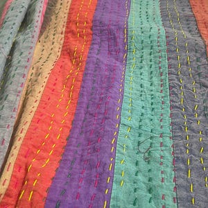 May include: A colorful, striped quilt with vertical bands of various colors, including red, purple, and teal. The quilt is hand-stitched with contrasting thread, adding texture and detail to the design. The overall aesthetic is a blend of rustic and vibrant.