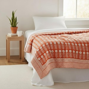 May include: A terracotta and white floral patterned quilt on a bed with white bedding. The quilt has a repeating floral design and a striped border. A small wooden side table with a plant and a mug is next to the bed.