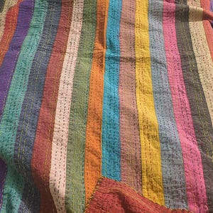 May include: A colorful, striped quilt with vertical bands of various colors, including red, blue, green, and yellow. The quilt has a stitched texture and a folded corner revealing a solid red fabric. The stitching is yellow.