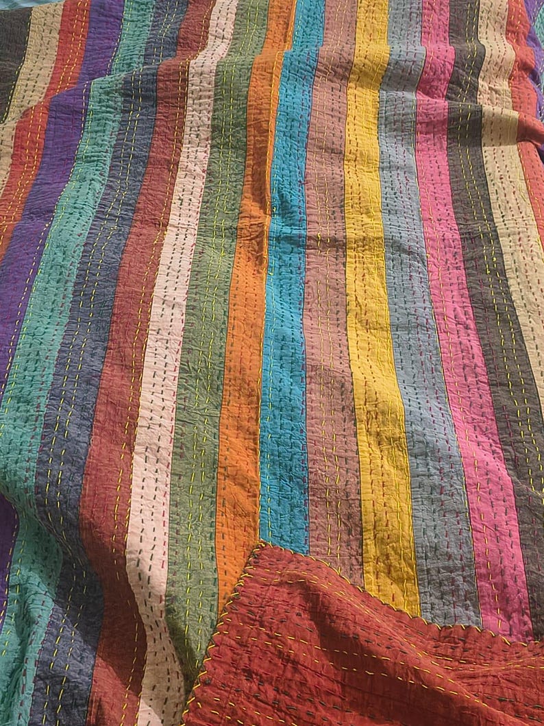 May include: A colorful, striped quilt with vertical bands of various colors, including red, blue, green, and yellow. The quilt has a stitched texture and a folded corner revealing a solid red fabric. The stitching is yellow.