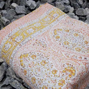 May include: Folded white and peach-colored textile with a yellow floral border and paisley pattern. The textile has a striped border in shades of peach and white. The design features yellow flowers and intricate details.