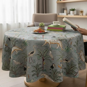 May include: A round tablecloth with a jungle-themed pattern. The gray fabric features illustrations of leopards, birds, and palm trees. The tablecloth is on a wooden table, with a plate of food and a book visible.