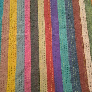 May include: A colorful textile with vertical stripes in various colors, including teal, yellow, red, and purple. The fabric has a textured appearance with visible stitching in a contrasting yellow thread, creating a patchwork effect. The overall design is a vibrant and eye-catching display.