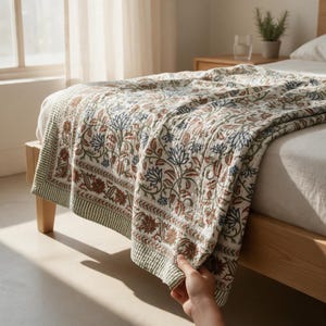 May include: A quilted bedspread featuring a repeating floral pattern in shades of rust orange, sage green, and dark blue on a cream background. The textile has a stitched texture and a wide border with a striped edge, draped over a wooden bed frame.