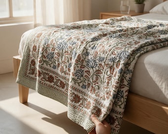 May include: A quilted bedspread featuring a repeating floral pattern in shades of rust orange, sage green, and dark blue on a cream background. The textile has a stitched texture and a wide border with a striped edge, draped over a wooden bed frame.